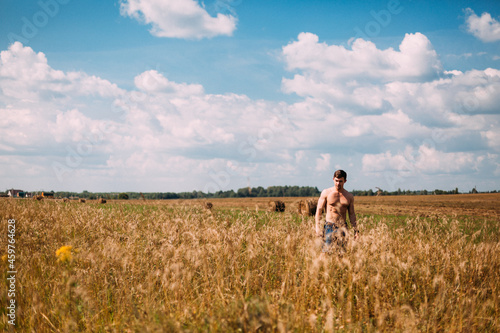 Naked to the torso, a man walks through a field of wheat. The sky is blue with white clouds. The man is wearing denim shorts. The field goes beyond the horizon. Lots of golden spikelets