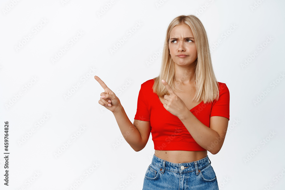 Sad grumpy blond girl pointing at upper left corner, frowning and looking uspet, standing against white background Sad grumpy blond girl pointing at upper left corner, frowning and looking uspet, standing against white background