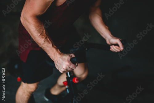 A man is doing a bike workout. In the frame there is a trunk, legs and a bicycle, close-up. Cycling training. A burgundy T-shirt and black shorts. Strong, sinewy arms, visible veins and tendons