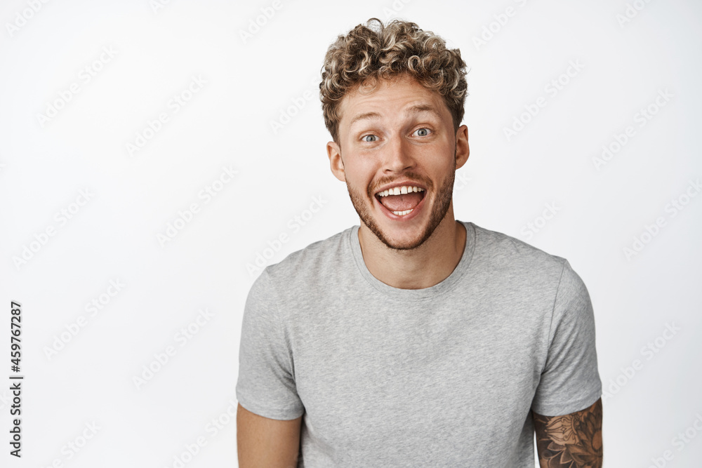 Image of excited blond guy smiling, looking amazed and happy at camera, hear big announcement, standing in gray t-shirt over white background Image of excited blond guy smiling, looking amazed and happy at camera, hear big announcement, standing in gray t-shirt over white background