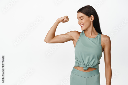 Smiling fitness girl shows strong arm, flexing biceps and look pleased, result of workout in gym, standing in activewear against white background