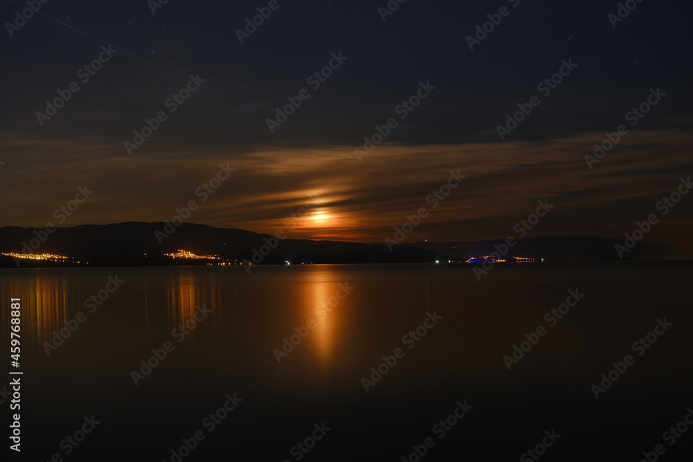 Full moon and moon reflection on water of iznik lake at middle of the night during bright night. Some village lights coming from hills. Full moon and moon reflection on water of iznik lake at middle of the night during bright night. Some village lights coming from hills.