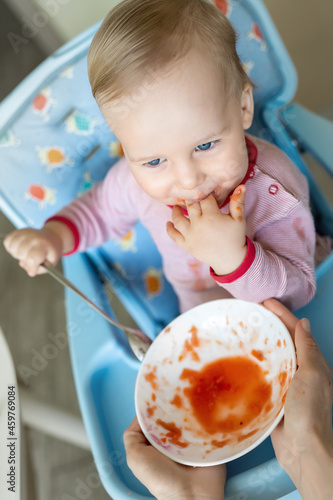 Cute happy adorable infant baby boy enjoy having fun sit in chair learning eat with spoon and plate healthy vegetable soup puree at home kitchen. Children feeding healthy food nutrition diet concept photo