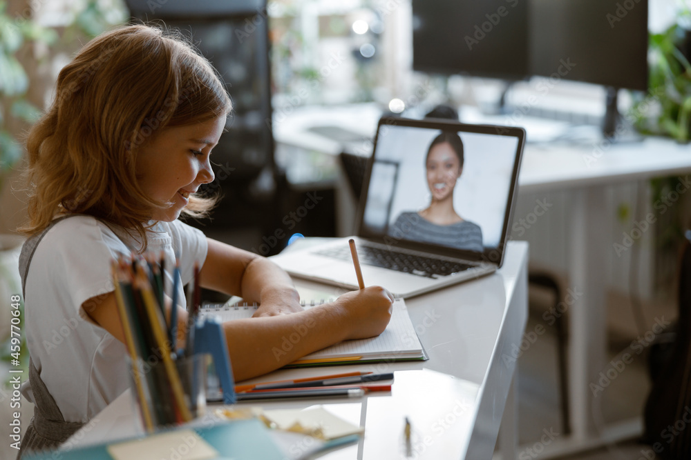 Smiling girl writes doing teacher task at video lesson via laptop at table in light room Smiling girl writes doing teacher task at video lesson via laptop at table in light room