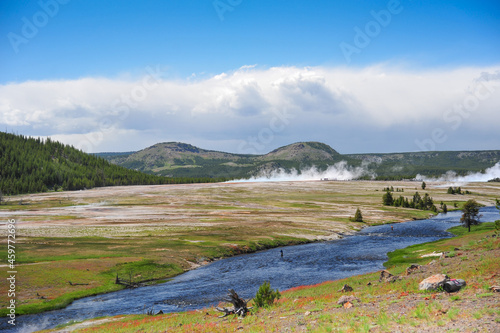 Yellowstone National Park as geyser blows hot water over the barren landscape as dark clouds roll in and fisherman fish the river 