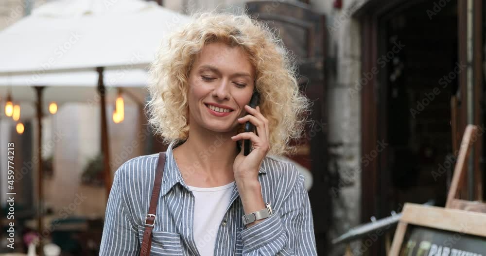 Waist up portrait view of the caucasian blonde curly woman chatting at the smartphone with somebody with pleasure smile while walking at the street Waist up portrait view of the caucasian blonde curly woman chatting at the smartphone with somebody with pleasure smile while walking at the street