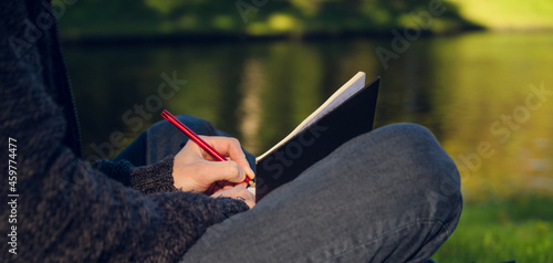 Student is sitting in the park on the grass and making notes. Pond on background.