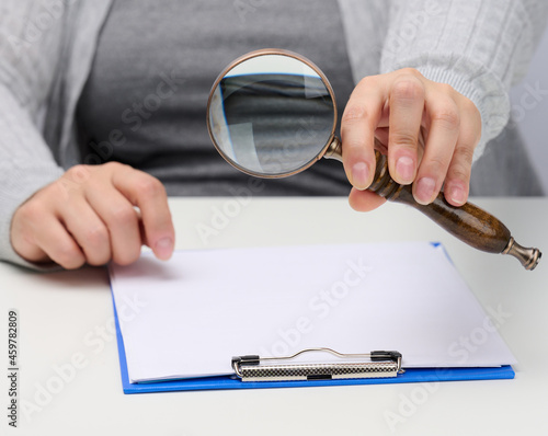 female hand holds a wooden magnifying glass over a white table with documents. Search for answers to questions, business analysis