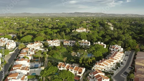 Overview of Quinta do Lago resort buildings in Almancil, Algarve, Portugal, Europe. Aerial shot of rooftops of luxury cottages in green landscape with mountains on background, 4k footage