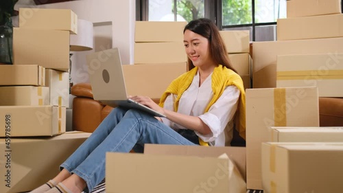 Asian woman with casual cloth sit among stack of boxes and use laptop with happiness in concept of small business to professional delivery product of online sell during new normal lifestyle.