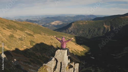 Woman rising up hands aerial. Tourist girl sit on mountain rock top. Nature landscape at autumn day. Mount hills with grass. Mountaineering lifestyle. Carpathians ridges, Ukraine, Europe. Drone shot