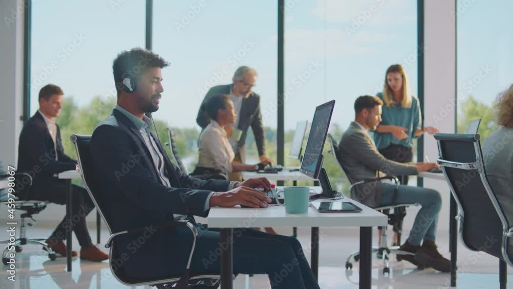 Mid adult business professional having online call with costumer. Colleagues discussing in background. Modern open space office interior. Mid adult business professional having online call with costumer. Colleagues discussing in background. Modern open space office interior.