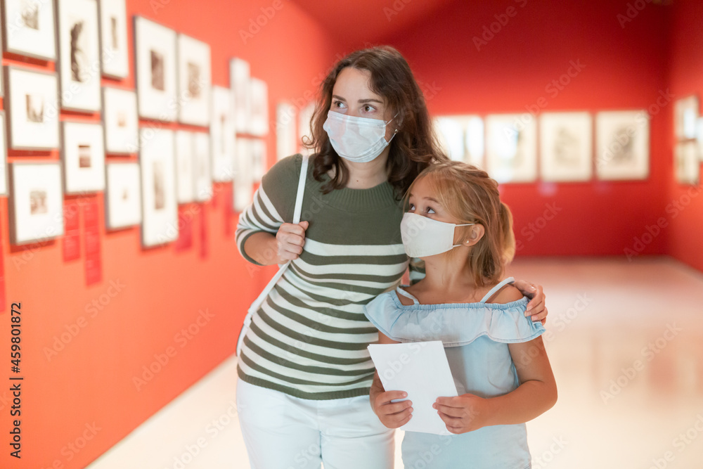 Portrait of woman with girl in medical masks standing in museum of art and looking at painting