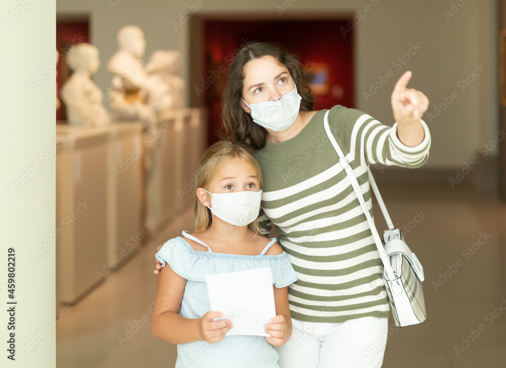 Woman with daughter in medical masks in museum of arts, pointing to art objects