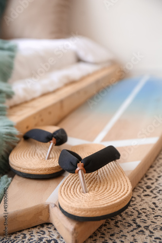 Wooden surfboard with stylish flip-flops on floor near bed