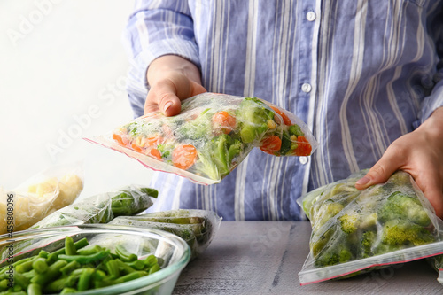 Woman holding plastic bags with frozen vegetables at table in kitchen, closeup