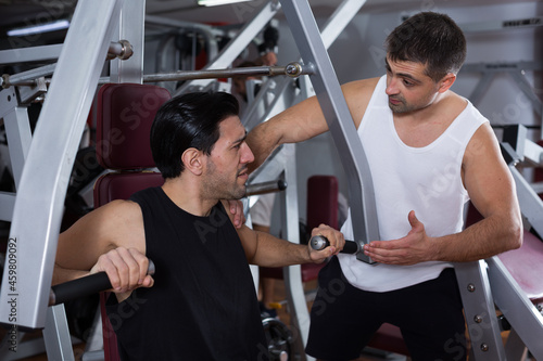 Athletic man helping friend to performing exercises on fitness machine in gym
