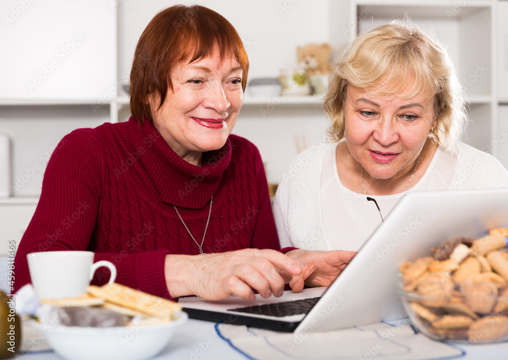 Smiling pensioners females talking at kitchen with laptop at home