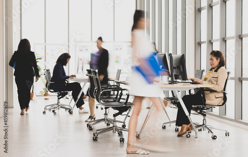 Selective focus on one business woman sitting and working on laptops on conference table with other blurry business women walking in rush in office with multiple glass windows