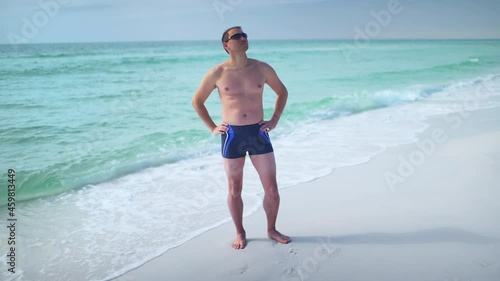 Panning one young man in swimming trunks with sunglasses looking at ocean turquoise water paradise in Seaside, Florida Gulf of Mexico beach on panhandle