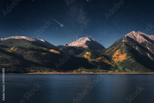 Nighttime scene along the shoreline of a lake with mountains in the background and a shooting star streaking across the sky. 