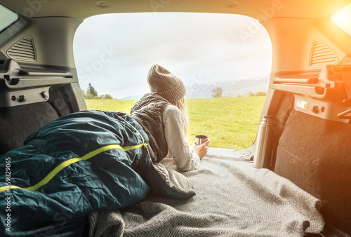 Girl resting in her car. Woman hiker, hiking backpacker traveler camper in sleeping bag, drinking hot tea and relaxing on top of mountain. Health care, authenticity, sense of balance and calmness.