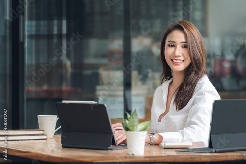 Fotografie Portrait of young beautiful asian woman smiling and looking at camera while sitting at her office desk