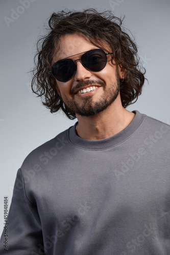 indoor portrait of handsome freckled smiling male with curly hair, wears sepctacles posing for social advertisement, isolated on gray wall with copy space for your promotional information