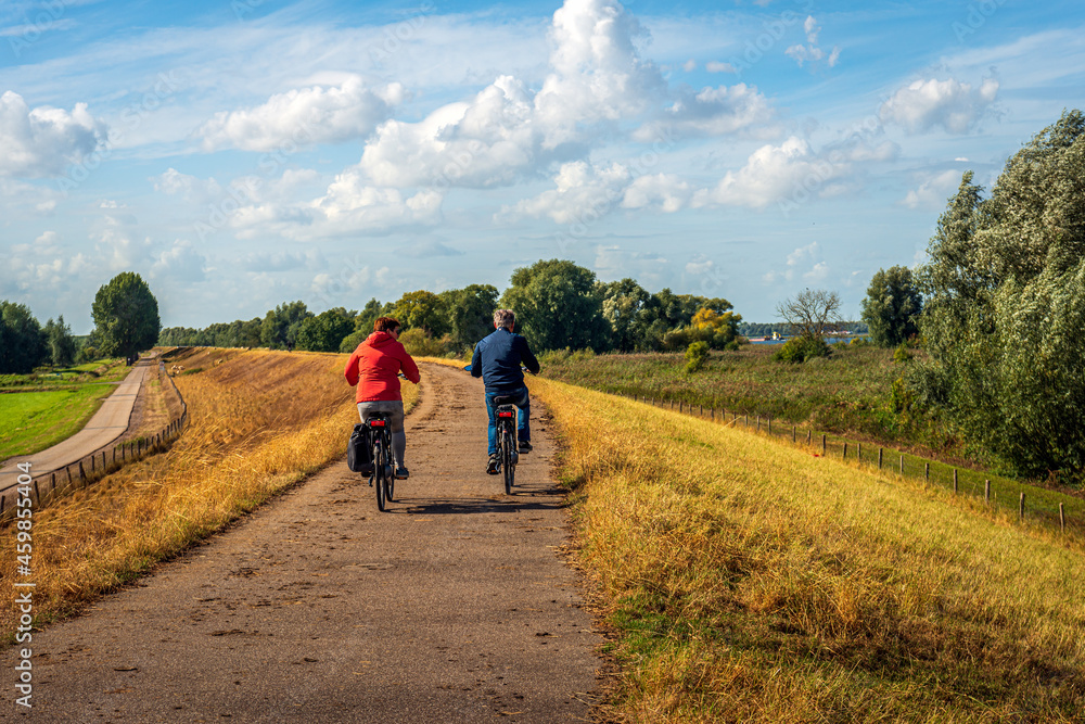 A man and woman cycle side by side on a Dutch dike. The grass on the slope of the dike has yellowed due to drought. It is a sunny day at the beginning of the autumn season.