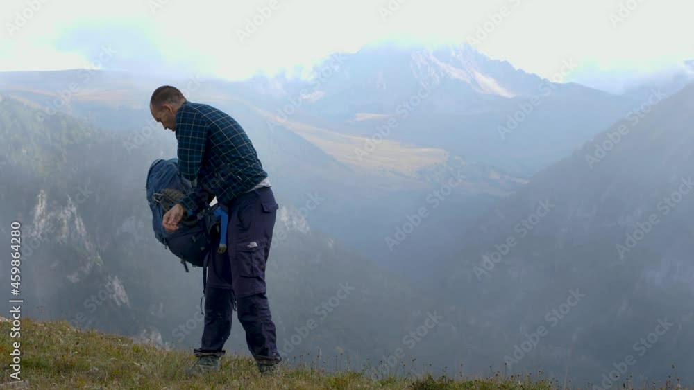 A male hiker in a plaid fleece jacket removes his backpack from his back. In the background is a gorge and snowy mountain tops. The concept of hiking A male hiker in a plaid fleece jacket removes his backpack from his back. In the background is a gorge and snowy mountain tops. The concept of hiking