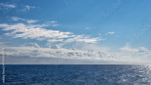 A front of cumulus clouds over a shiny sea, horizontally panoramic.
