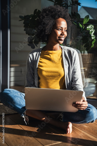Smiling woman with laptop sitting cross-legged in sunlight at home