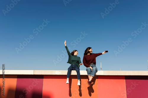 Carefree female friends taking selfie while sitting on retaining wall