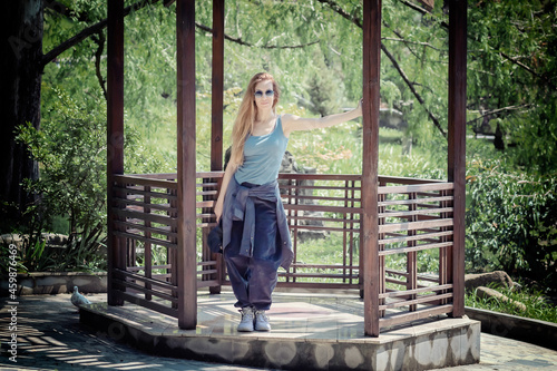 Long-haired woman in hip-hop outfit stands in a gazebo holding a wooden beam in a green summer park