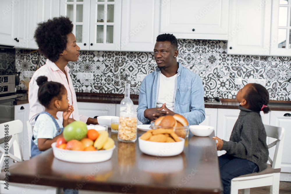African man and woman sitting on modern kitchen with two cute girls eating, drinking and chatting. Happy family spending morning time together at home. Healthy nutrition and parenting concept.