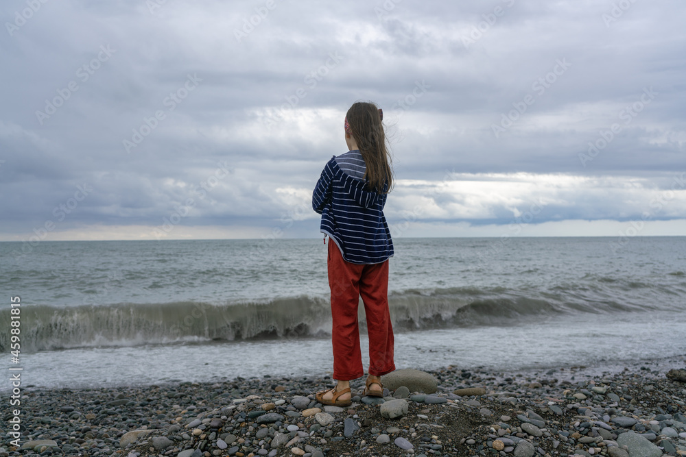 A little girl stands on the seashore near the sea waves on the beach during a storm and rain