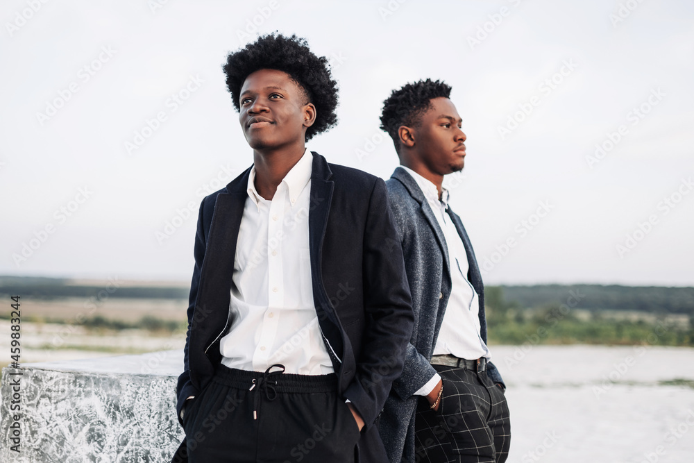 Two handsome black men posing in elegant suits, looking to the side, posing against the backdrop of rocky mountains Two handsome black men posing in elegant suits, looking to the side, posing against the backdrop of rocky mountains