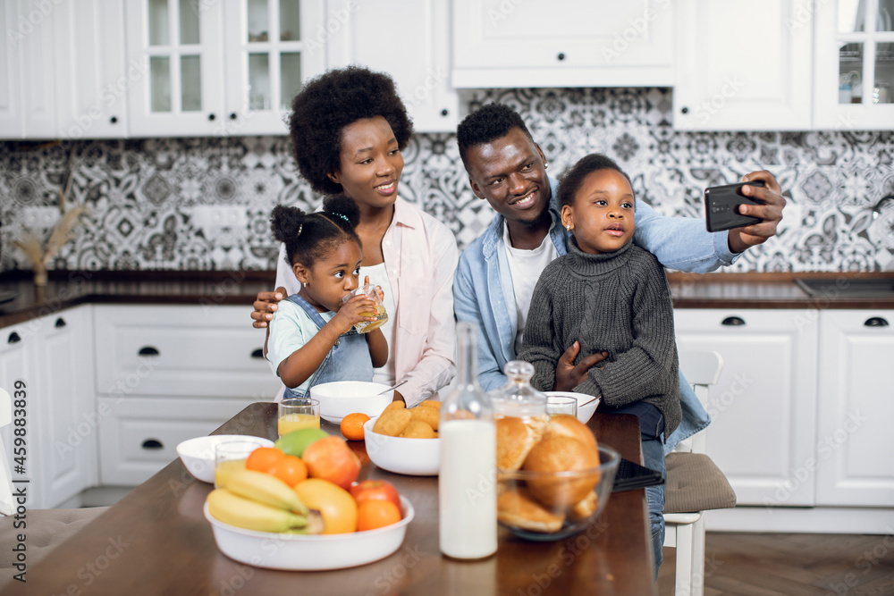 Positive african family of four taking selfies on modern smartphone while having tasty breakfast on bright kitchen. Concept of morning time, home and technology.