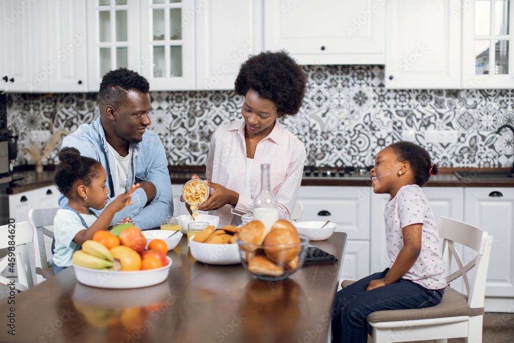 African american family of four eating cereals with milk during morning time at home. Young parents with two daughters having breakfast together on modern kitchen.