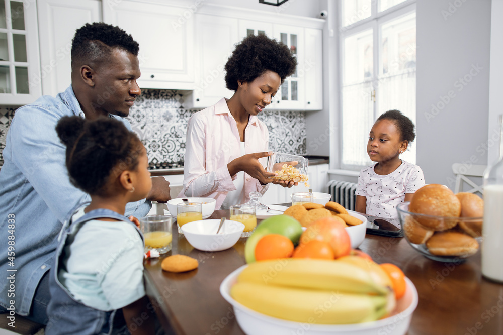 Pretty african sisters sitting with young parents on modern bright kitchen. Positive family eating cereals with milk for breakfast. Morning time at home. Pretty african sisters sitting with young parents on modern bright kitchen. Positive family eating cereals with milk for breakfast. Morning time at home.