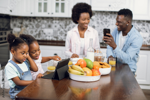 African american parents sitting on kitchen and using smartphone while their cute daughters watching cartoon on digital tablet. Modern gadgets for lifestyles. Morning time at home.