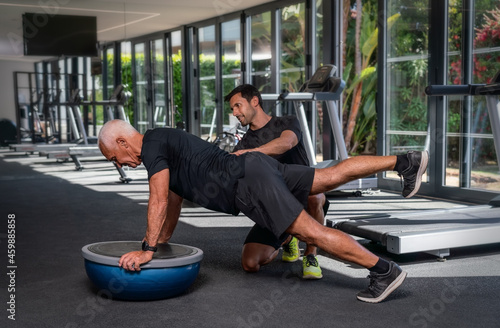 Fotografija An elderly Caucasian man, in the gym, with a personal fitness trainer, in rehabilitation, doing exercises for balance on a gymnastic ball