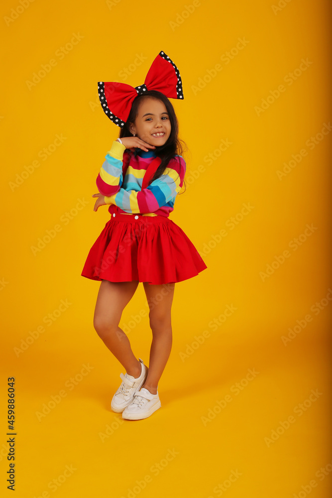 a dark-skinned happy girl in a red skirt in a colored jacket with a red polka dot bow on her head on a yellow background a dark-skinned happy girl in a red skirt in a colored jacket with a red polka dot bow on her head on a yellow background