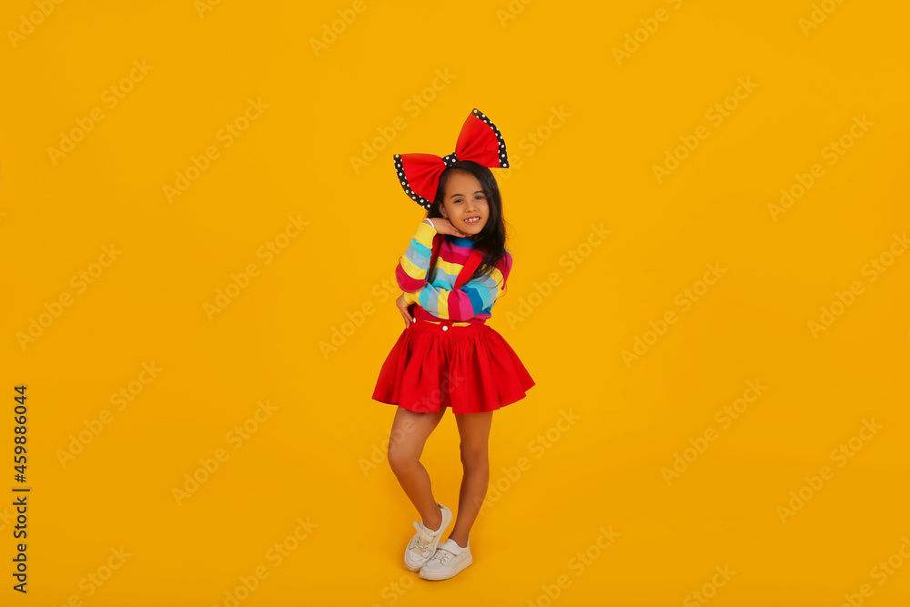 a dark-skinned happy girl in a red skirt in a colored jacket with a red polka dot bow on her head on a yellow background a dark-skinned happy girl in a red skirt in a colored jacket with a red polka dot bow on her head on a yellow background