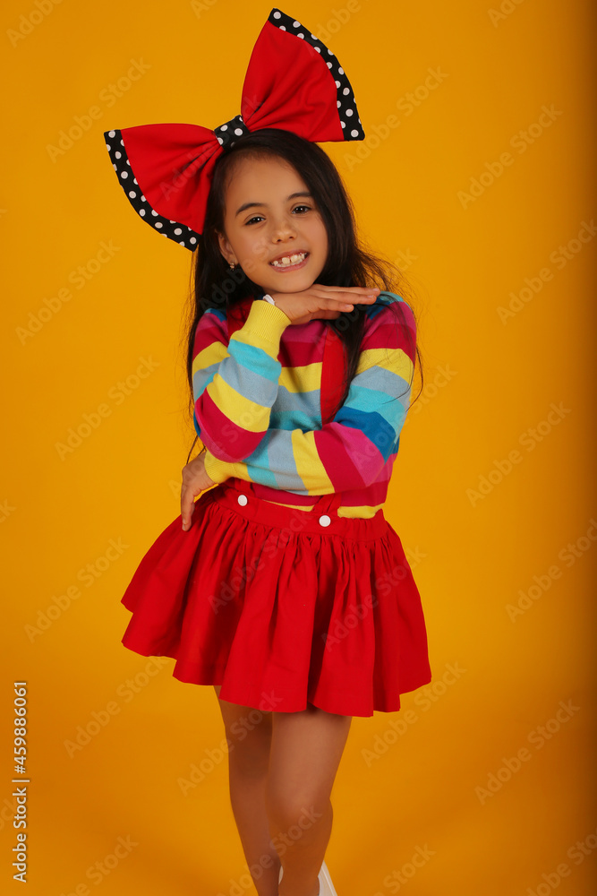 a dark-skinned happy girl in a red skirt in a colored jacket with a red polka dot bow on her head on a yellow background a dark-skinned happy girl in a red skirt in a colored jacket with a red polka dot bow on her head on a yellow background