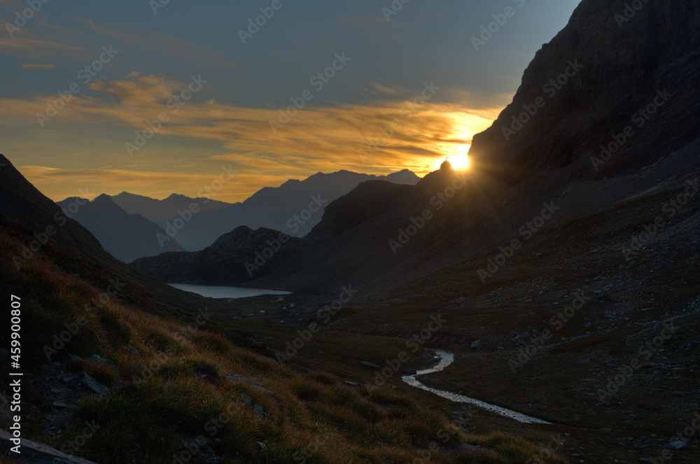 Early morning in the Alps: silhouettes of mountains in shades of hazy colors, a silvery lake and river in the foreground