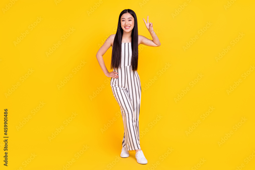 Full length body size photo girl smiling in striped overall showing v-sign gesture isolated vibrant yellow color background Full length body size photo girl smiling in striped overall showing v-sign gesture isolated vibrant yellow color background