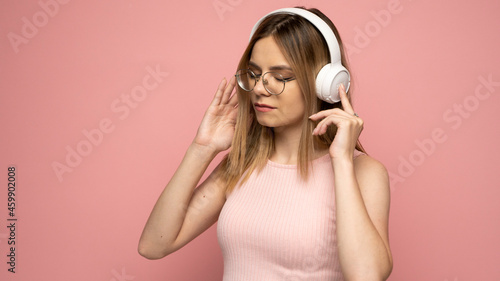 Beautiful attractive young blond woman wearing pink t-shirt and glasses in white headphones listening music and smiling on pink background in studio. Relaxing and enjoying. Lifestyle.