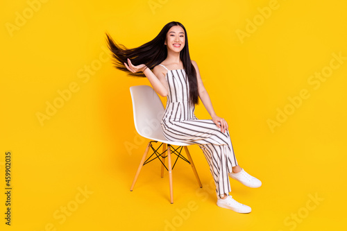 Full length body size photo young girl sitting on chair wearing casual overall smiling throwing hair isolated vivid yellow color background