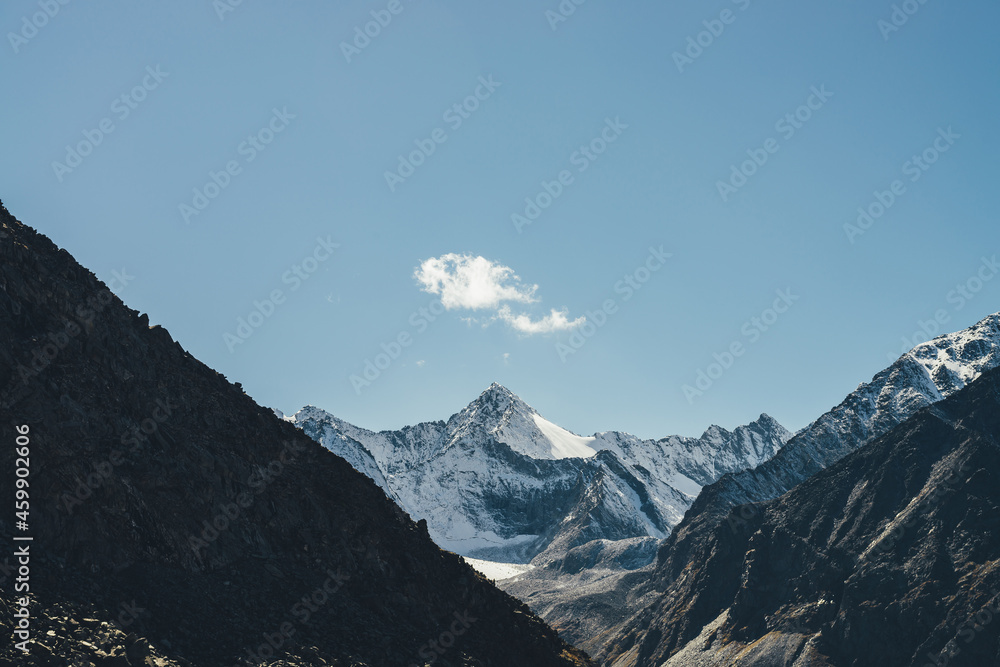 Alpine landscape with high snowy mountain with peaked top under cirrus clouds in sky. Atmospheric view to big snow covered mountains in sunshine. Black rocks and white-snow pointy peak in sunlight.